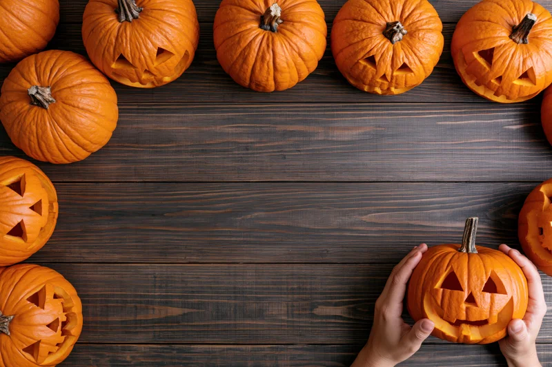 Hands holding a carved pumpkin on a wooden table surrounded by jack-o'-lanterns in a flat lay arrangement