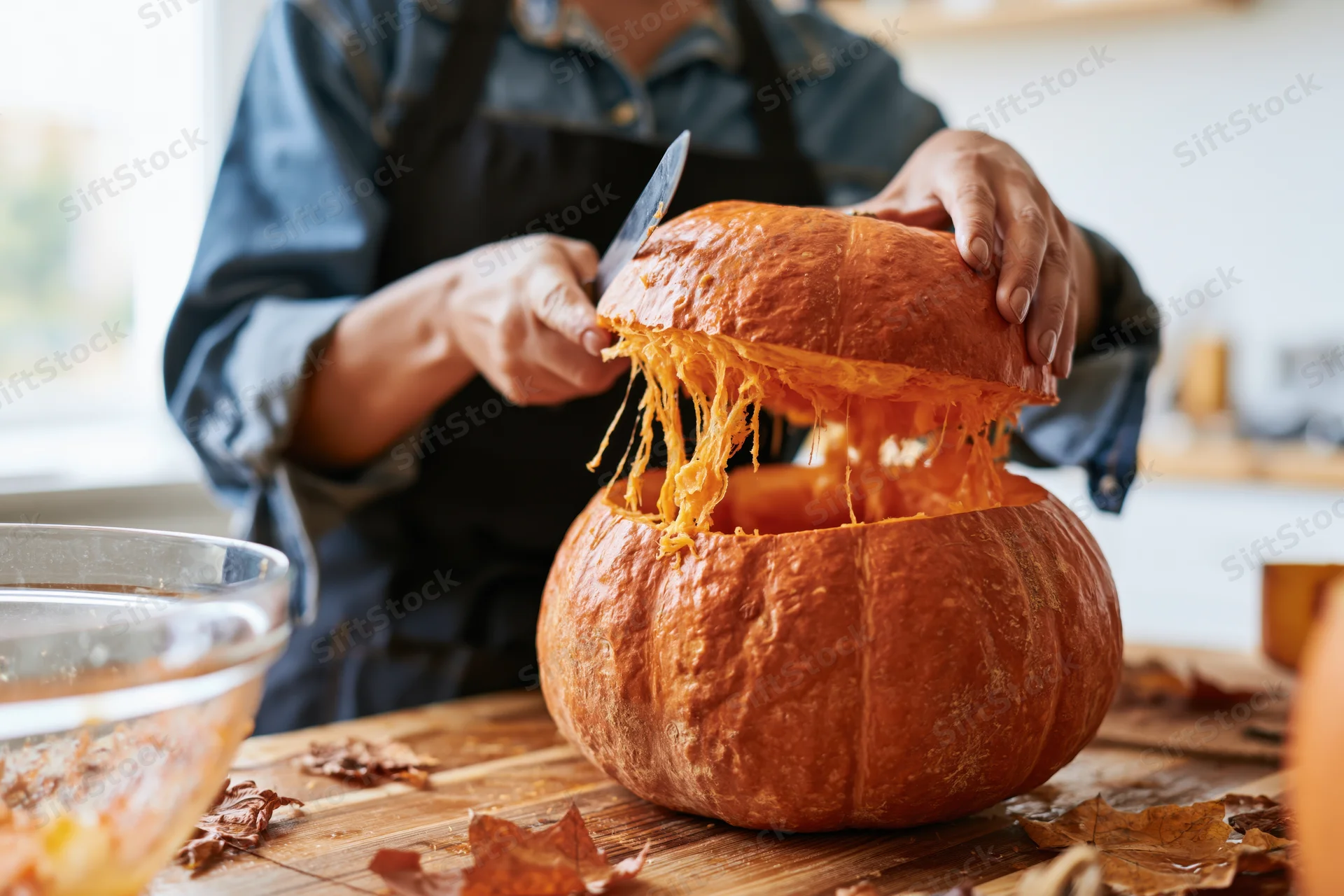 Woman carving a pumpkin on a wooden kitchen table surrounded by autumn leaves
