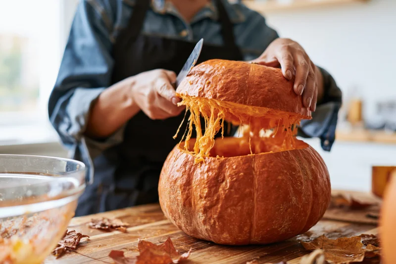 Woman carving a pumpkin on a wooden kitchen table surrounded by autumn leaves
