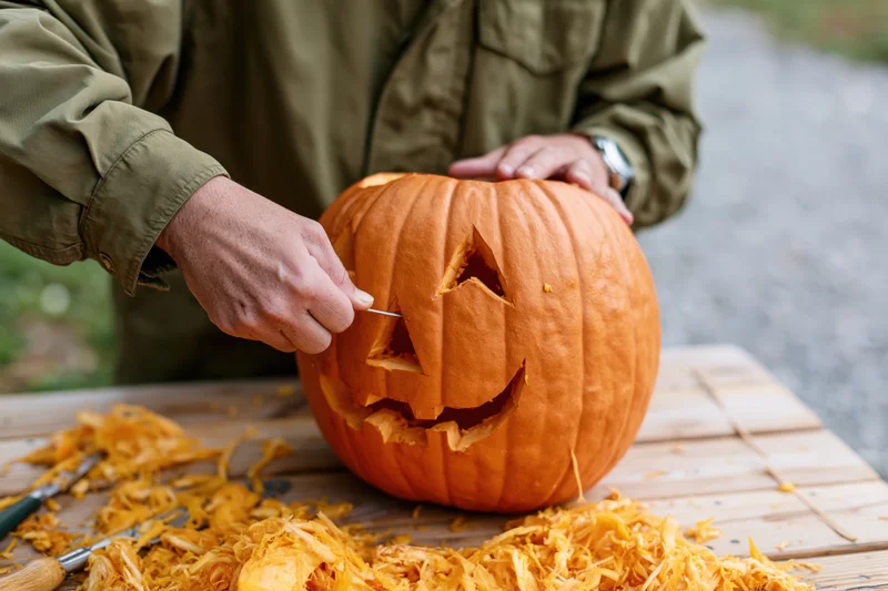Man carving a jack-o'-lantern face into a large orange pumpkin on a wooden table outdoors