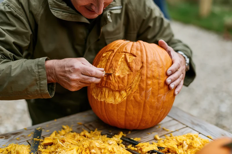 Man carving a detailed face into a large orange pumpkin on a wooden table outdoors