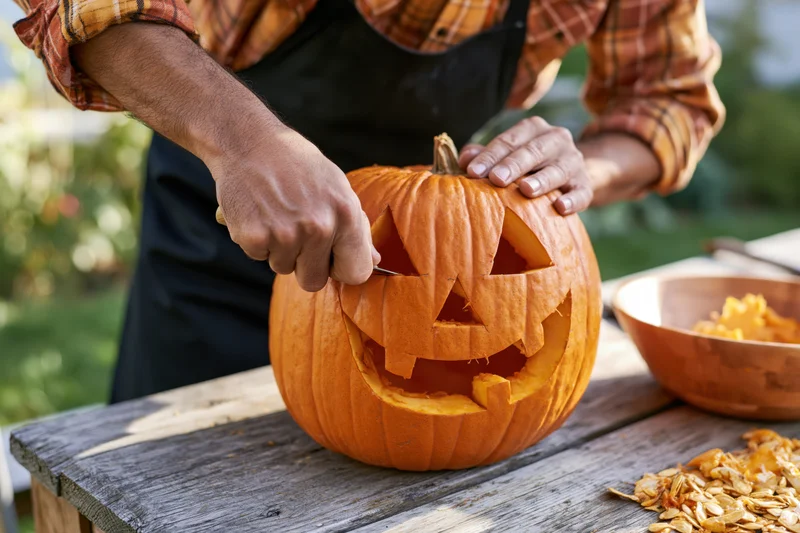 Man carving a jack-o-lantern face into a pumpkin on a wooden table outdoors