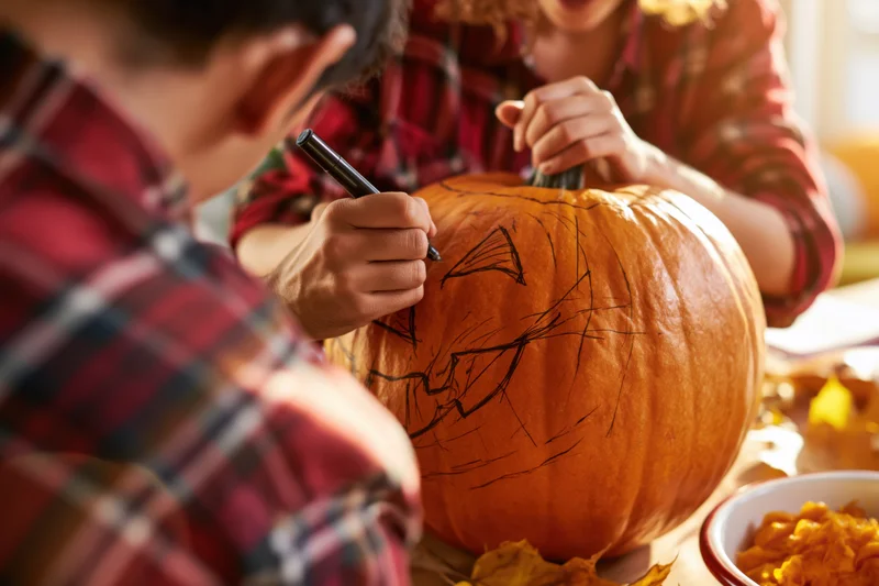 Person drawing jack-o-lantern face on large orange pumpkin at table