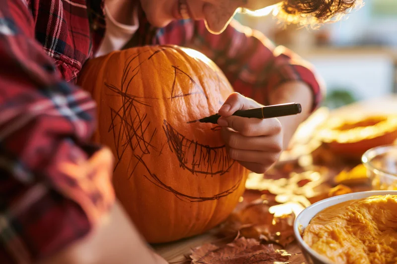 Young man drawing a jack-o-lantern face on a pumpkin at a kitchen table