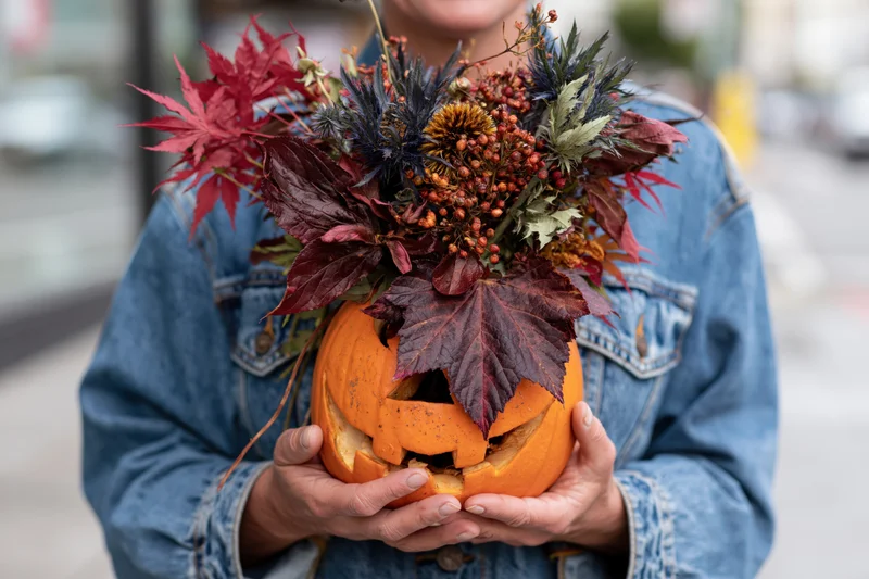 Woman holding carved jack-o-lantern filled with autumn floral arrangement outdoors