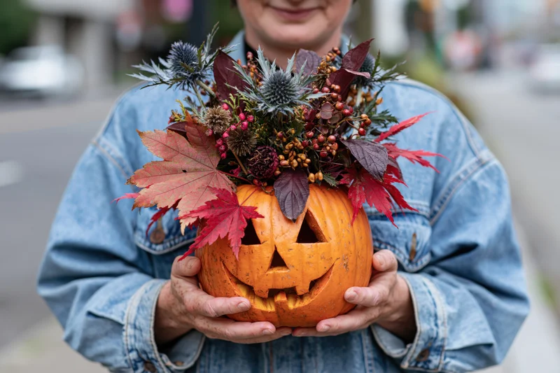 Woman holding carved jack-o-lantern with floral arrangement in denim jacket