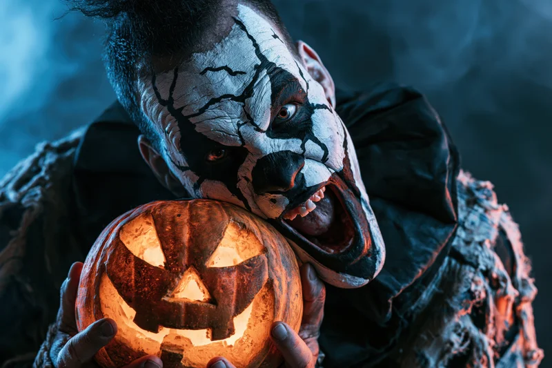 Male clown with cracked white facepaint holding a glowing carved pumpkin in a smoky dark setting