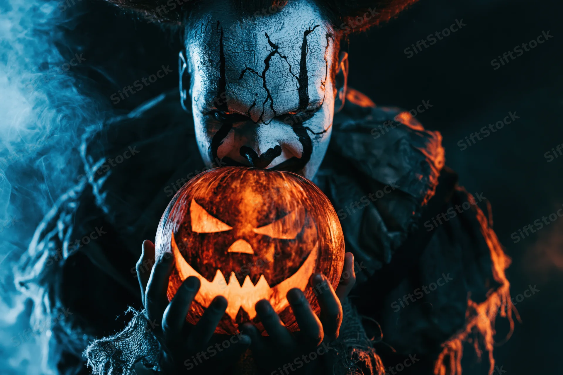 Creepy clown in Halloween costume holding glowing carved pumpkin in dark smoky setting