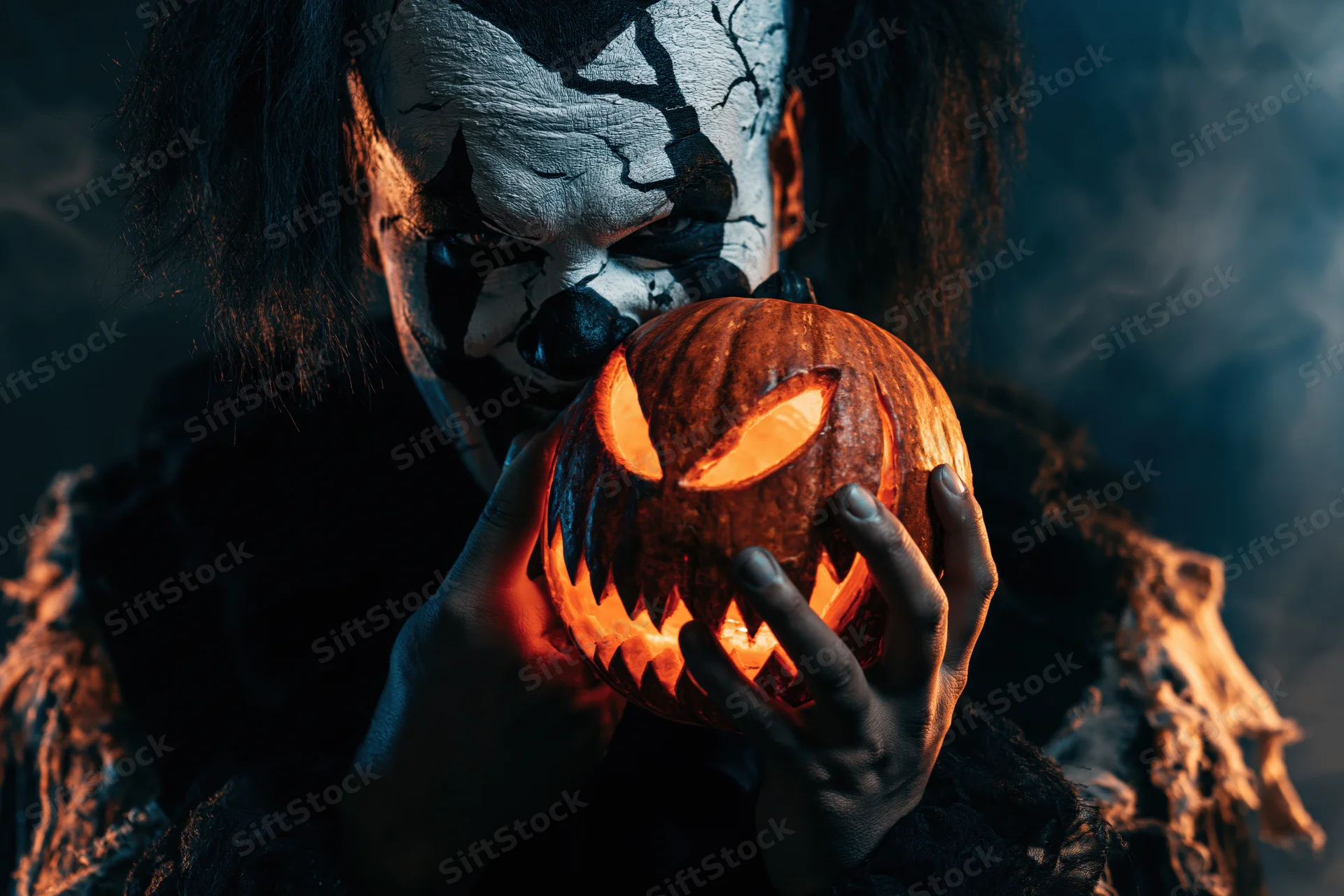 Creepy clown with cracked white makeup holding a glowing carved pumpkin in dark smoky setting
