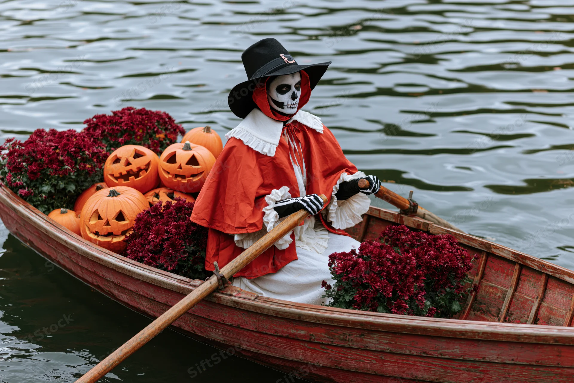 Person in skeleton face paint and red cape rowing a boat filled with pumpkins and flowers on water