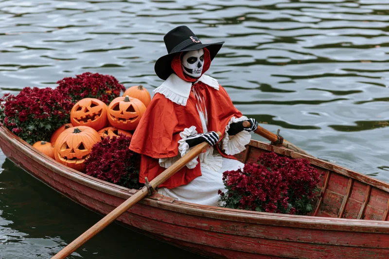 Person in skeleton face paint and red cape rowing a boat filled with pumpkins and flowers on water
