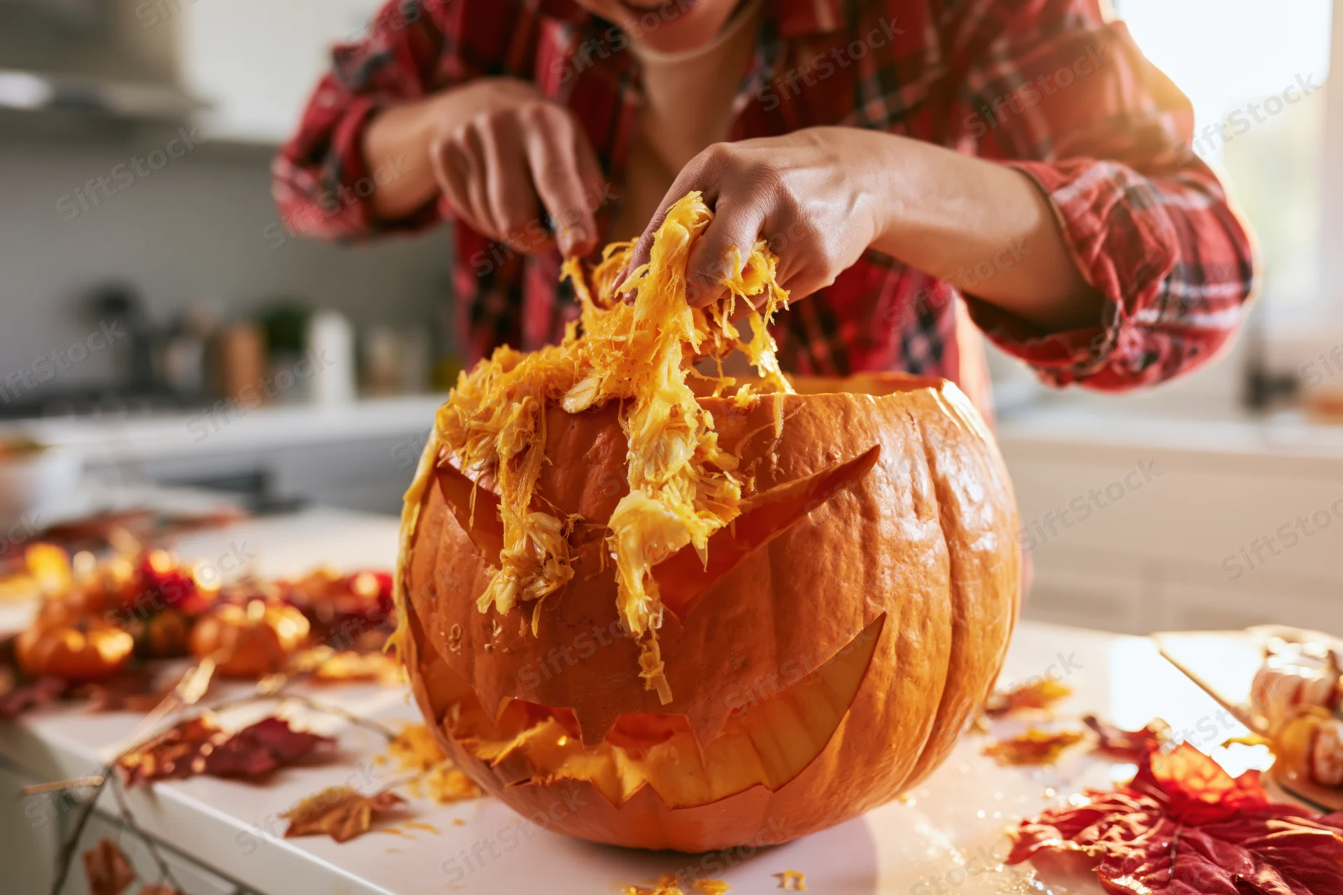 Woman scooping fibrous pumpkin innards from carved jack-o-lantern on kitchen countertop