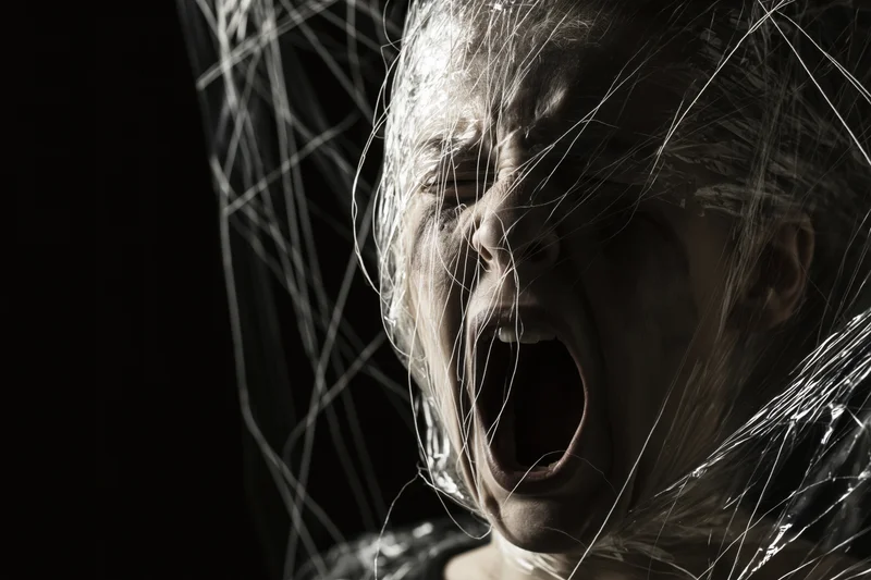 Woman screaming with mouth wide open, face partially wrapped in plastic sheeting, dark background