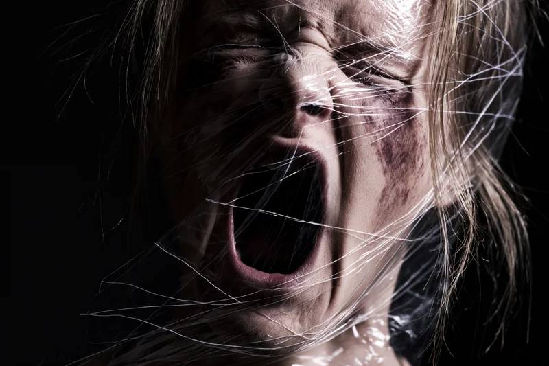 Woman screaming with mouth wide open, face partially wrapped in transparent plastic, dark background