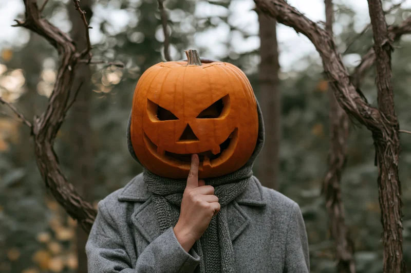 Person in gray coat with carved jack-o-lantern head covering finger to lips in forest