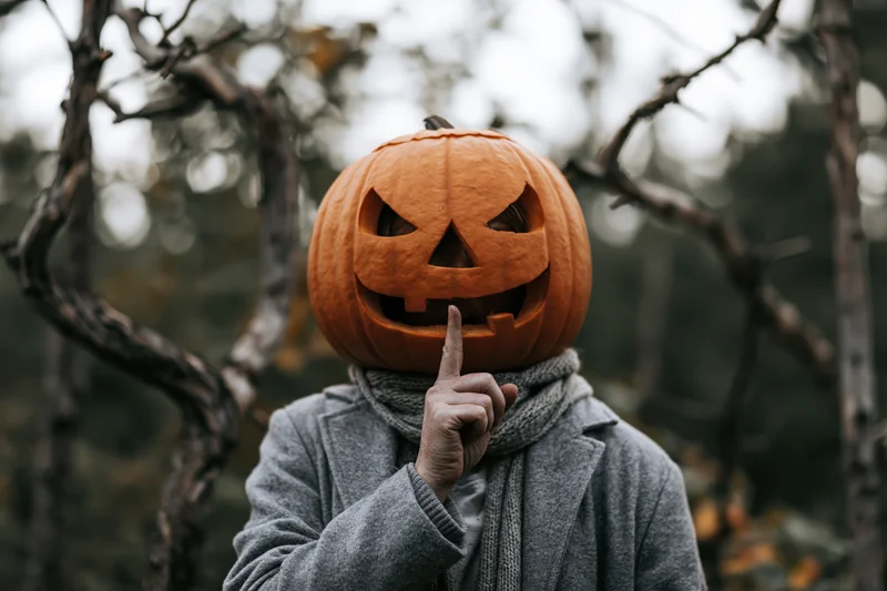 Person wearing carved jack-o-lantern over head making silence gesture in forest
