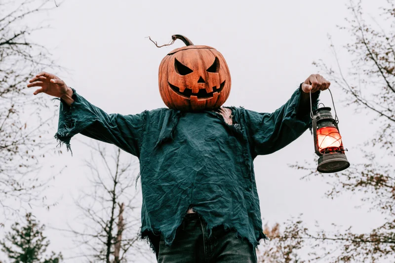 Person in carved pumpkin head mask holding lantern with arms outstretched in forest