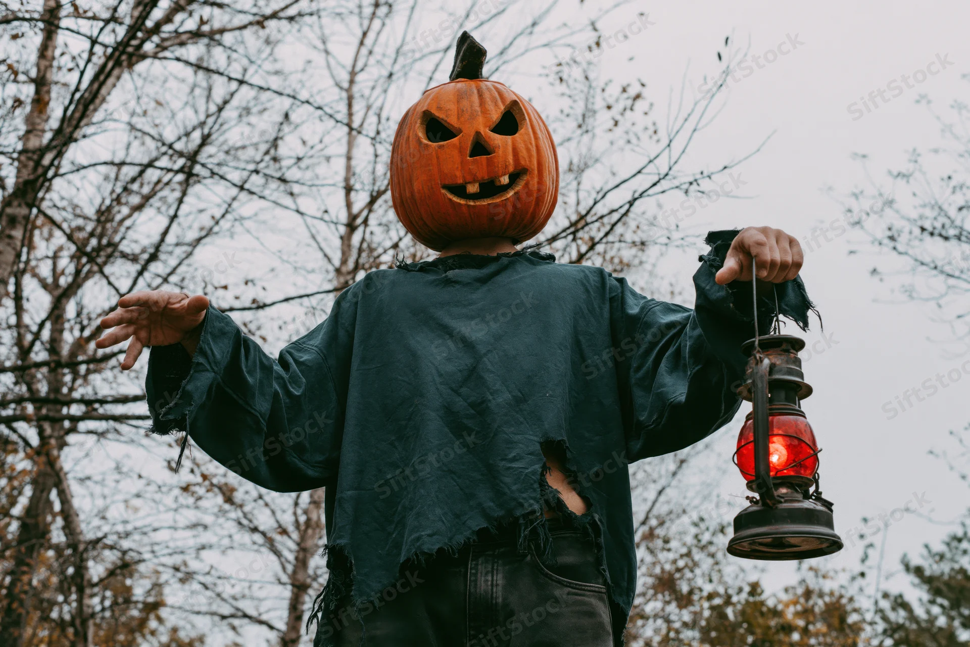 Person in carved pumpkin mask holding red lantern outdoors in autumn forest