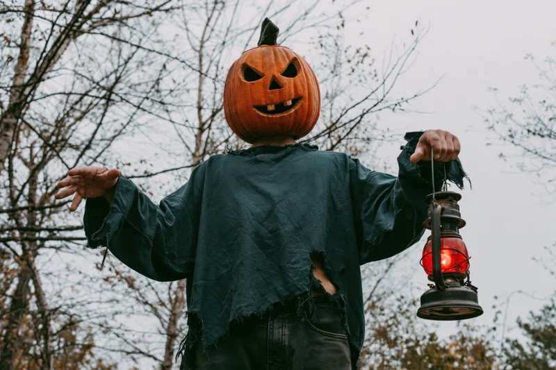 Person in carved pumpkin mask holding red lantern outdoors in autumn forest