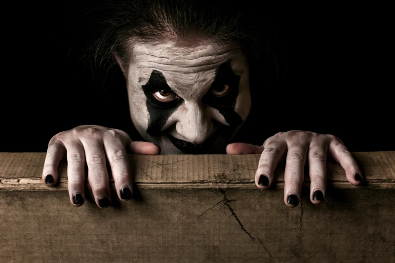 Clown with dark theatrical makeup peering over a cracked wooden board