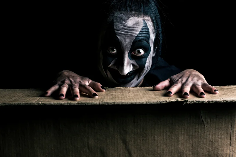 Clown with dark theatrical face paint peeking over a wooden surface in shadow