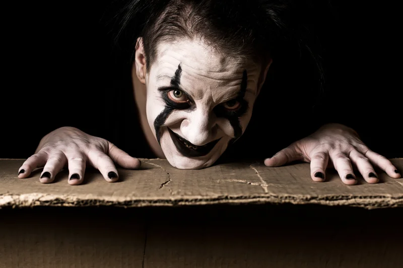 Male clown with dark theatrical makeup peering over cardboard box edge