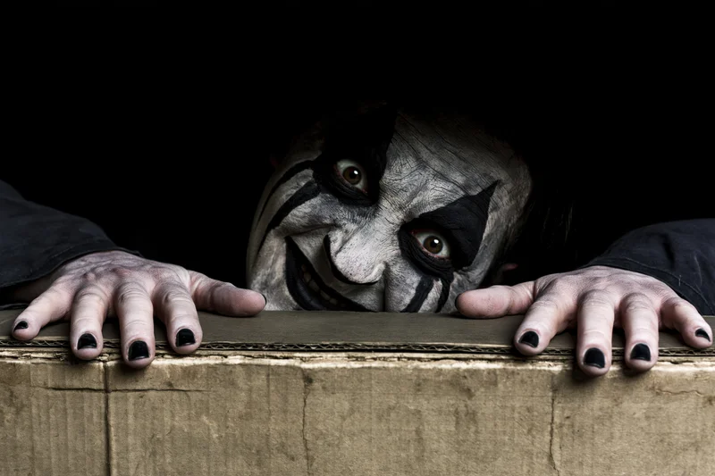 Clown with dark face paint peeking over cardboard box edge