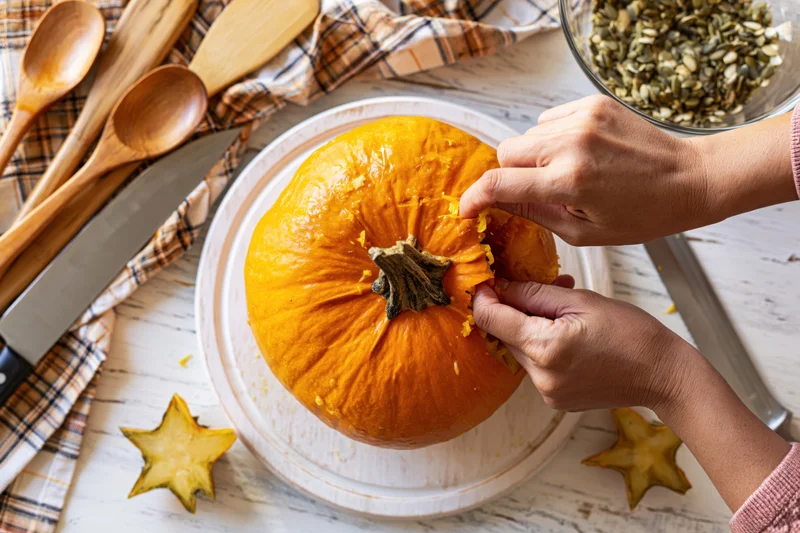 Hands carving a bright orange pumpkin on a round wooden cutting board in a kitchen setting