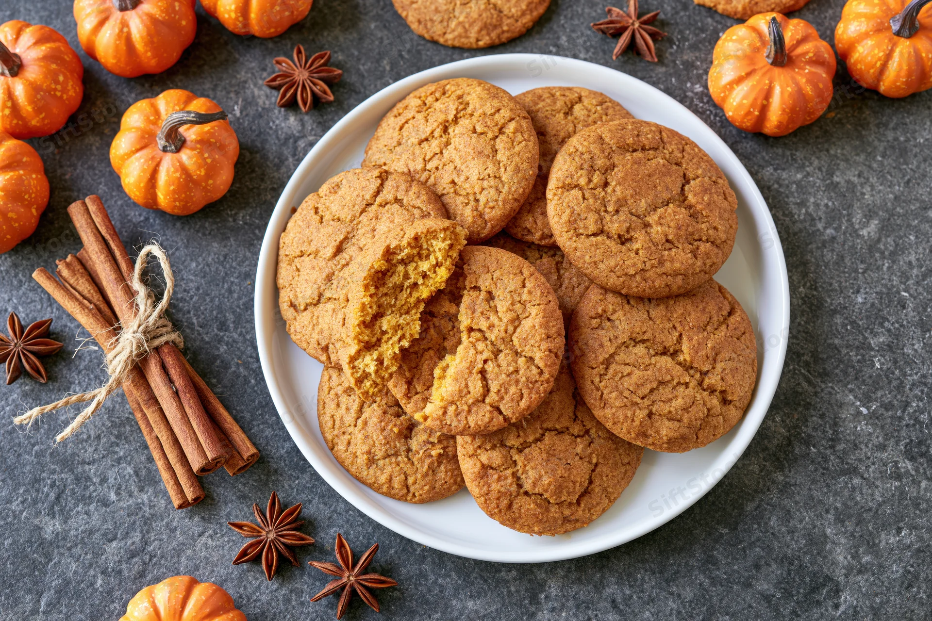 Pumpkin spice cookies piled on a white plate with one broken open, surrounded by small pumpkins, cinnamon sticks and star anise on a dark stone surface