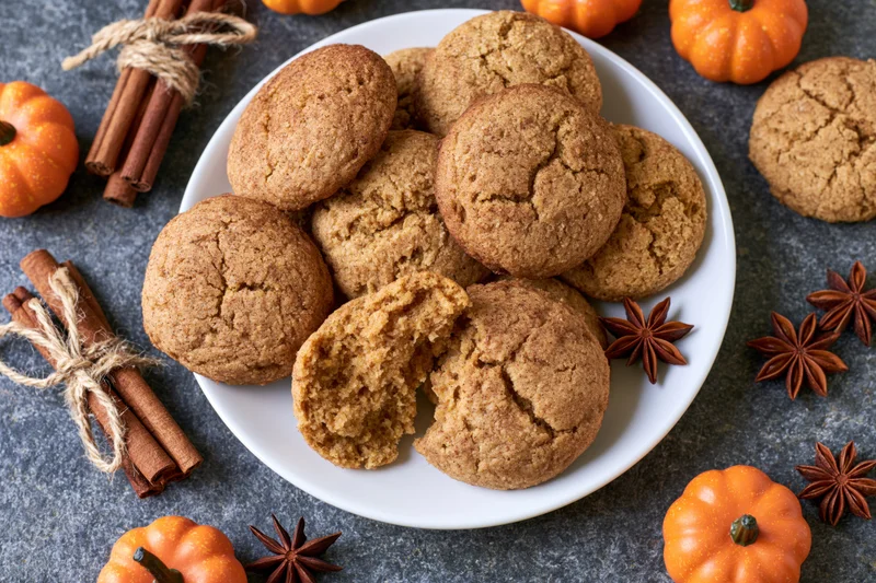 Pumpkin spice cookies piled on a white plate with one broken open, surrounded by small pumpkins, cinnamon sticks and star anise on a dark stone surface