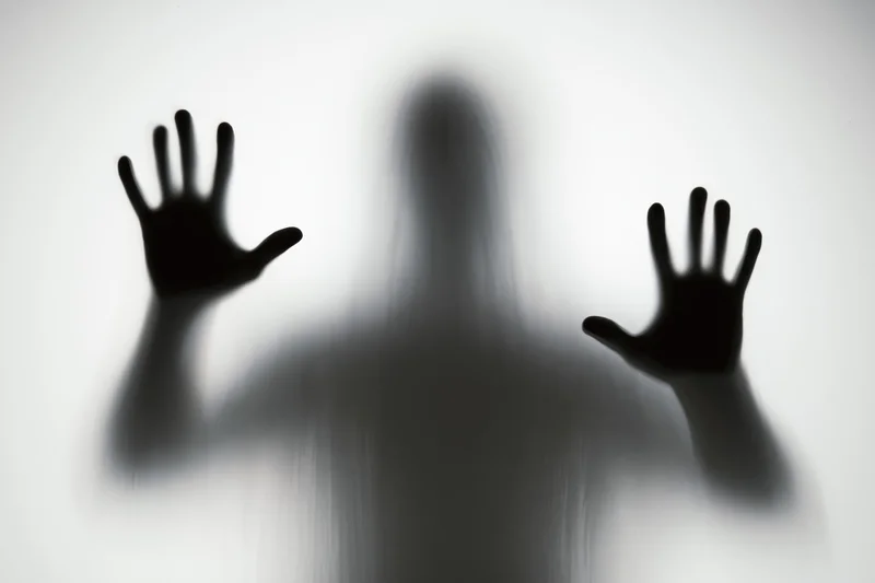 Shadowy silhouette of a person pressing hands flat against frosted glass