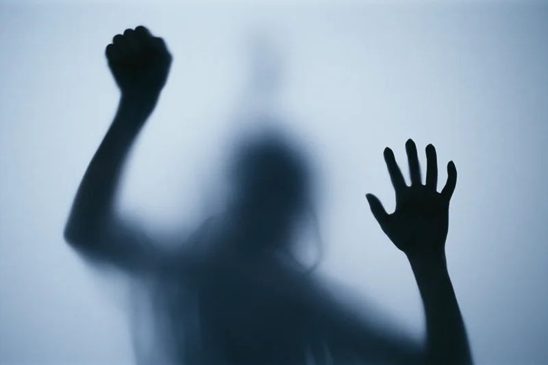 Silhouette of a woman pressing one hand flat against frosted glass with the other raised in a fist