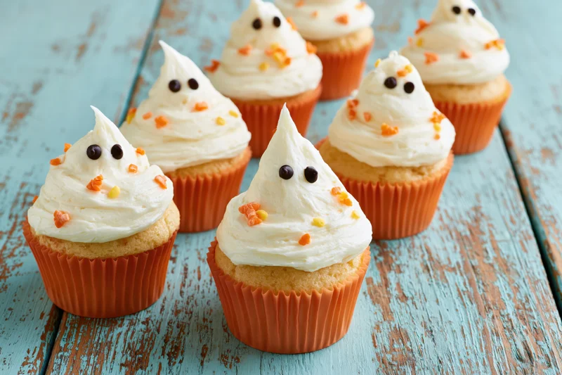 Ghost-themed cupcakes with white frosting and candy eyes arranged on a weathered blue wooden table