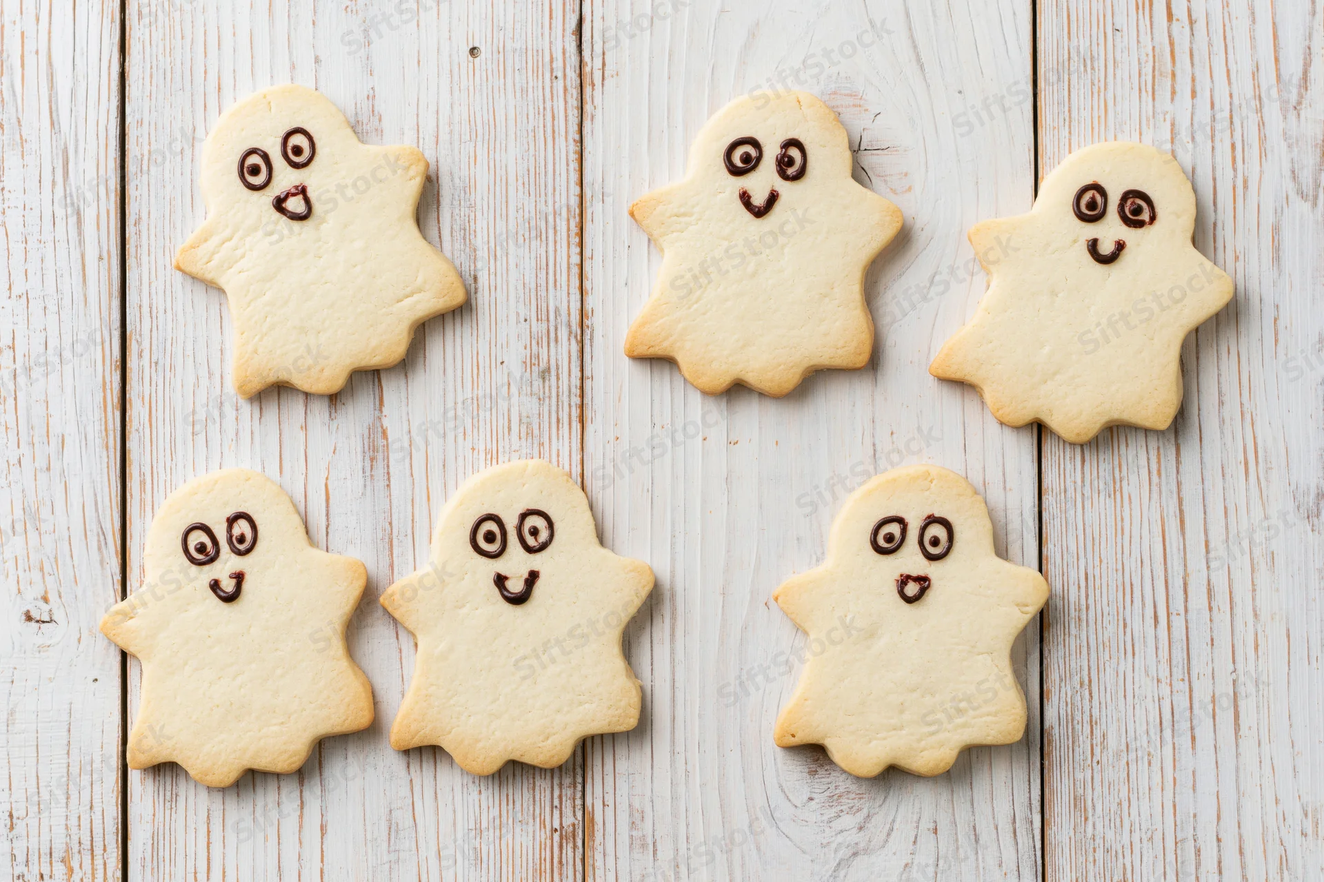 Six ghost-shaped shortbread cookies with chocolate icing faces arranged on a white wooden surface