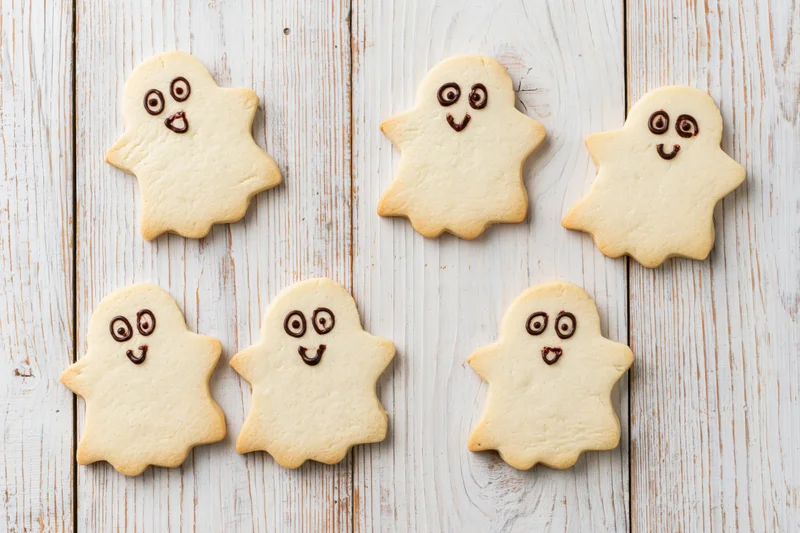 Six ghost-shaped shortbread cookies with chocolate icing faces arranged on a white wooden surface