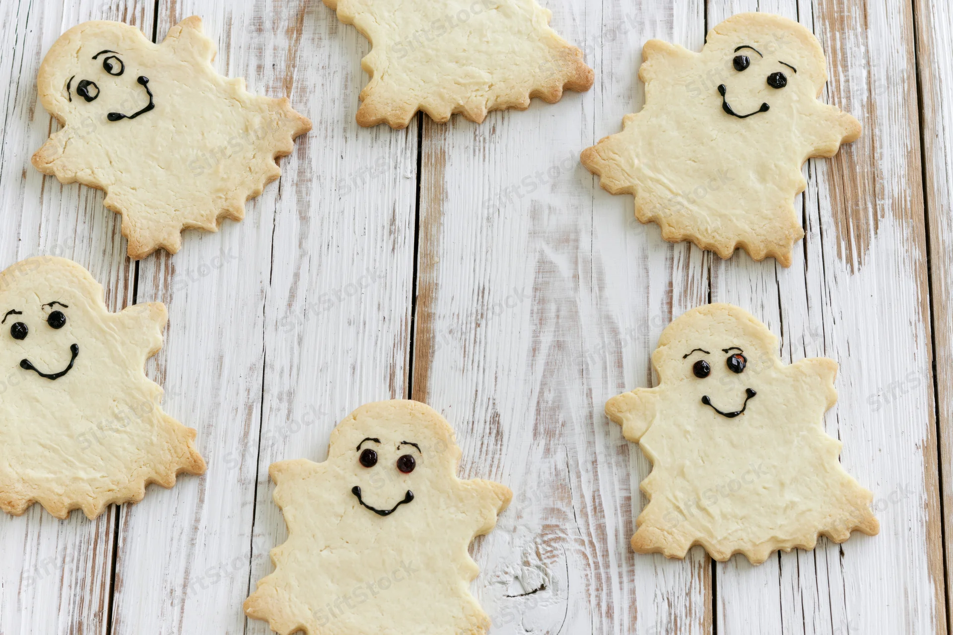 Ghost-shaped shortbread cookies with hand-drawn faces arranged on a white wooden surface