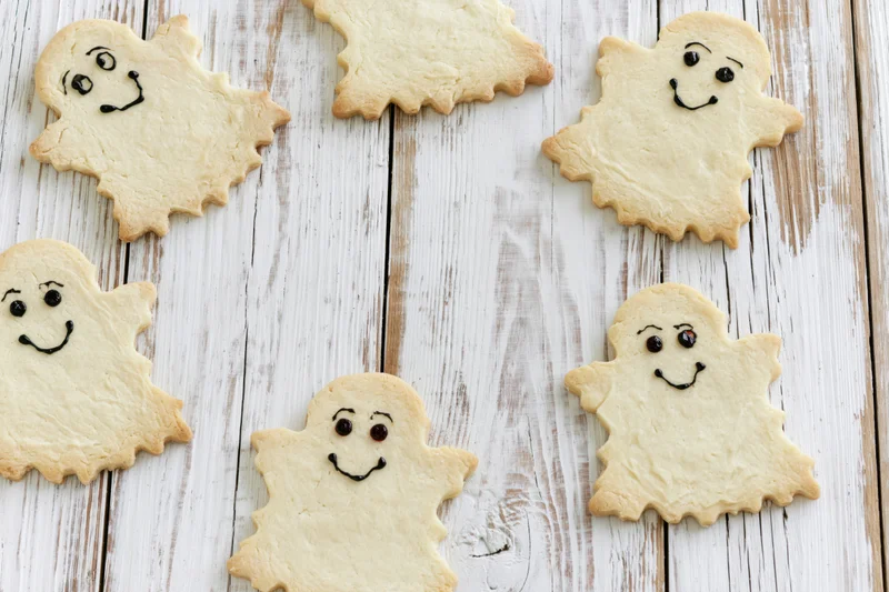 Ghost-shaped shortbread cookies with hand-drawn faces arranged on a white wooden surface