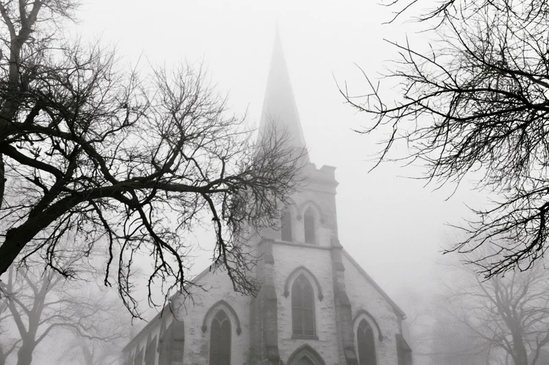 Gothic revival church with pointed spire seen through bare tree branches in fog