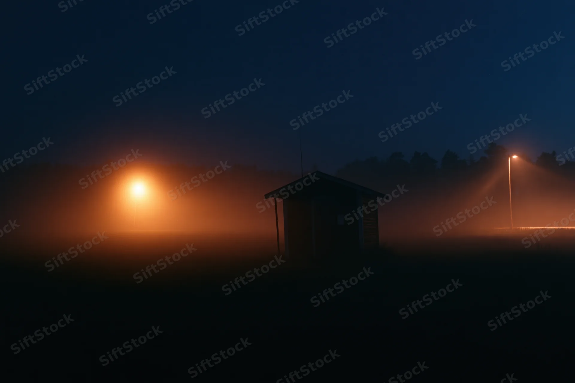 Small wooden shed silhouetted in fog on a rural field at night with glowing lights in the distance