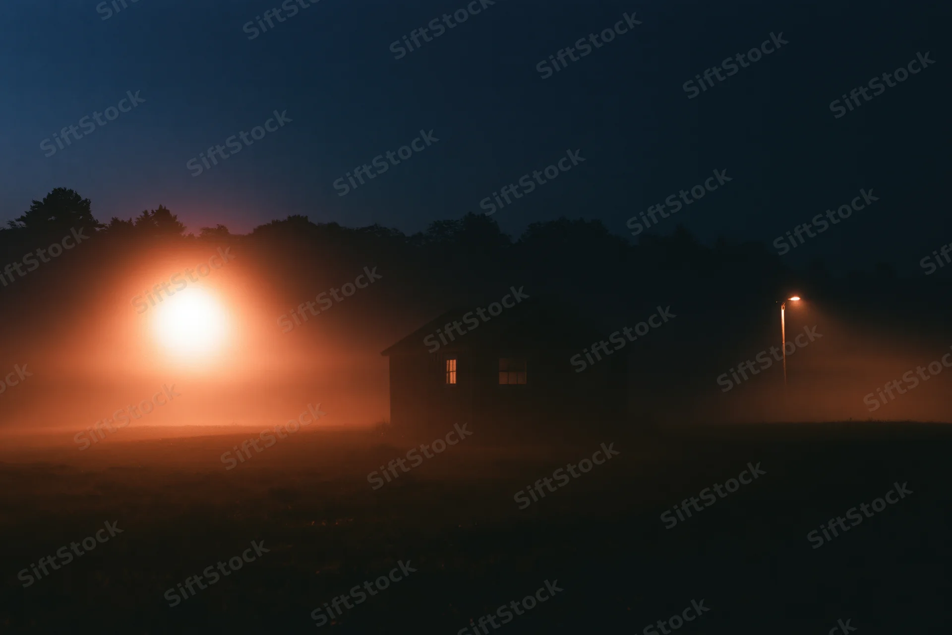 Small wooden shed silhouetted in fog on a rural field at night with warm glow from lamp and window light
