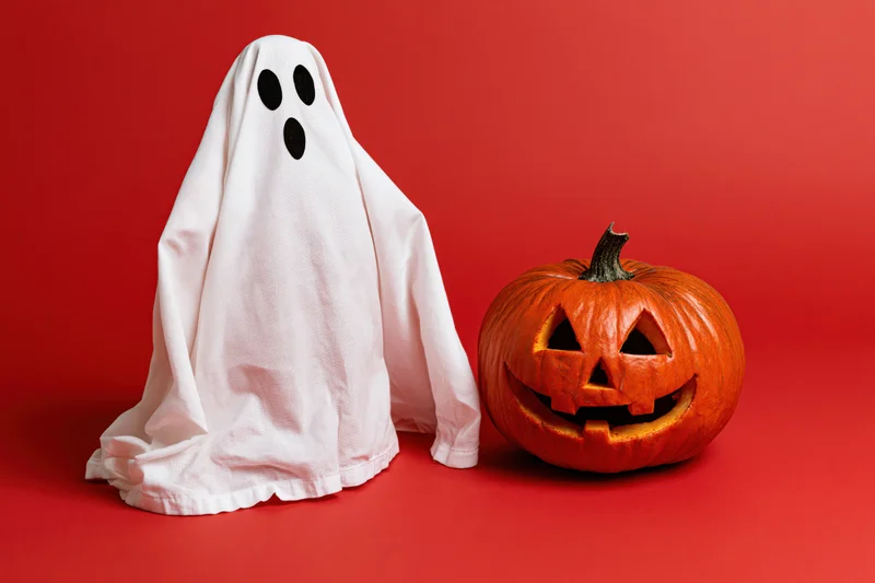 White fabric ghost costume beside carved pumpkin on red background