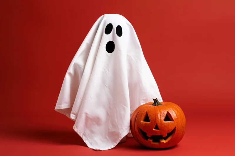 White fabric ghost costume with black eyes beside carved orange pumpkin on red background