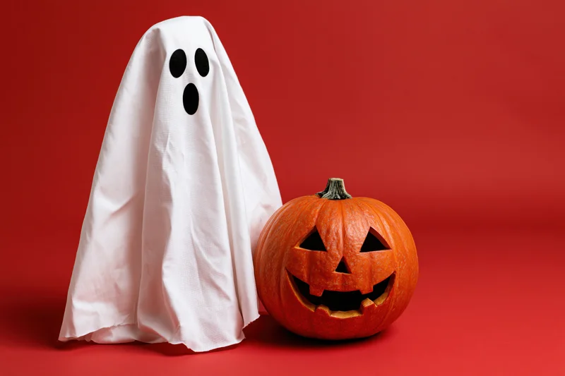 White fabric ghost costume beside carved pumpkin on red background