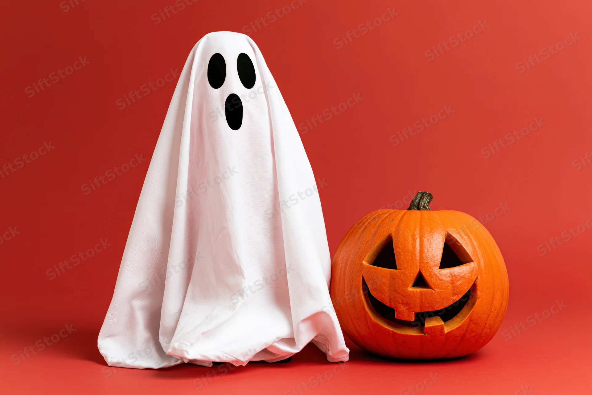 White fabric ghost costume with black eyes beside carved orange pumpkin on red background