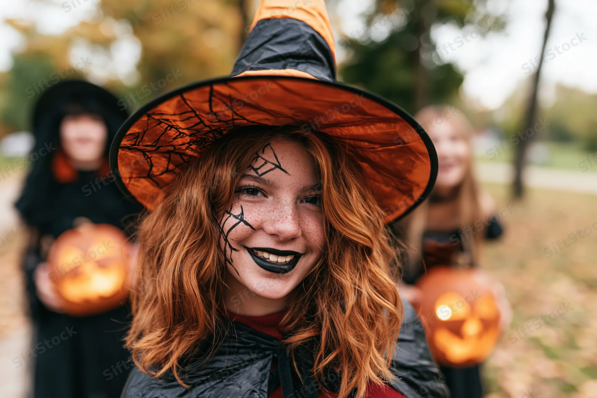 Teenage girl in witch costume with spiderweb hat and face paint smiling outdoors with friends holding jack-o-lanterns in autumn park