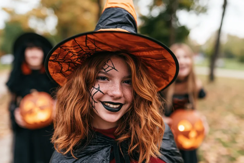 Teenage girl in witch costume with spiderweb hat and face paint smiling outdoors with friends holding jack-o-lanterns in autumn park