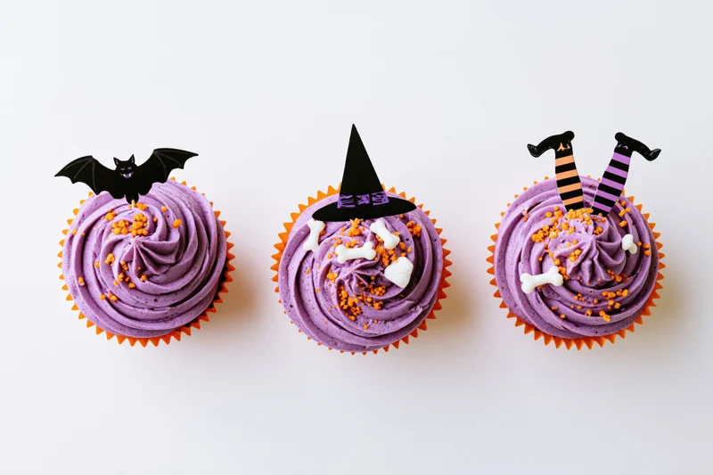 Three halloween-themed cupcakes with purple frosting and candy decorations arranged in a row on white background
