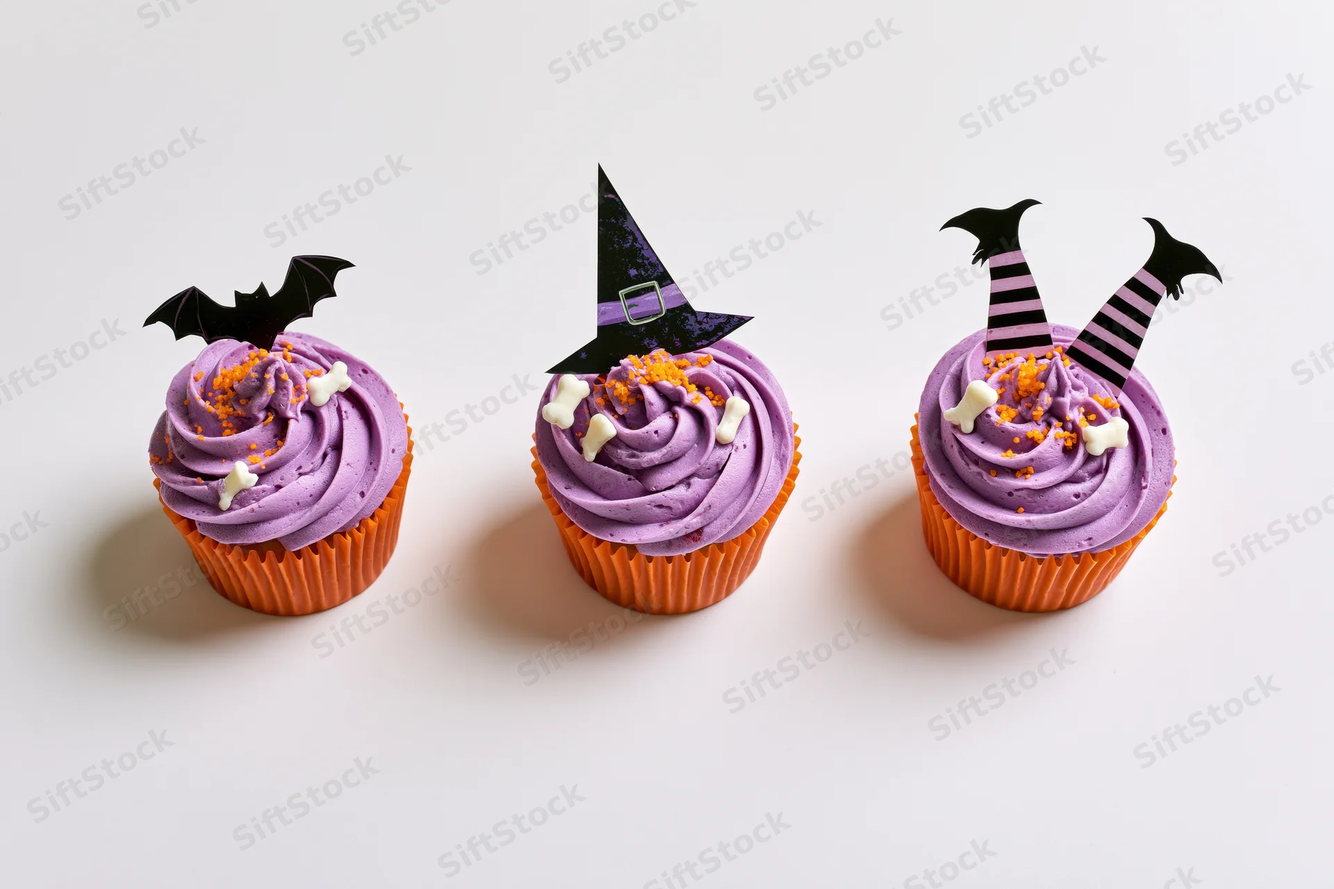 Three halloween-themed cupcakes with purple frosting and witch, bat, and legs decorations in orange wrappers on white background