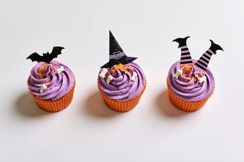Three halloween-themed cupcakes with purple frosting and witch, bat, and legs decorations in orange wrappers on white background
