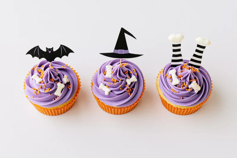 Three halloween-themed cupcakes with purple frosting and decorative toppers arranged in a row on a plain white surface